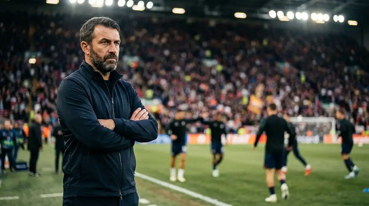 A tactical close-up of a football coach on the touchline in a dark tracksuit, arms folded, watching intently under stadium lights, players warming up out of focus behind him